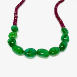 Può includere: Una collana con grandi perle di smeraldo ovali di un verde vibrante. Gli smeraldi sono intervallati da perline più piccole, rotonde e rosso intenso, creando un contrasto sorprendente. La collana è esposta su uno sfondo bianco.