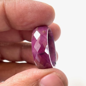 Puede incluir: Un anillo de rubí facetado. El anillo es de color magenta intenso con múltiples facetas que captan la luz. El diseño del anillo es una banda sólida con un interior pulido. El anillo se sostiene entre los dedos.