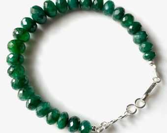 May include: A beaded bracelet featuring faceted, deep green gemstones. The bracelet is strung together with a silver-colored clasp. The gemstones are oval-shaped and have a polished, reflective surface. The bracelet is displayed on a white surface.
