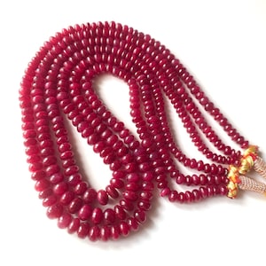 Könnte beinhalten: Eine mehrsträngige Halskette aus kleinen, runden, tiefroten Perlen. Die Perlen sind miteinander aufgereiht und erzeugen einen mehrschichtigen Effekt. Die Halskette hat einen goldfarbenen Verschluss und wird vor weißem Hintergrund präsentiert.