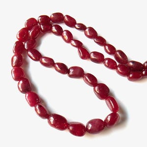 Puede incluir: Un collar compuesto por piedras preciosas pulidas, de forma ovalada y de color rojo intenso. Las cuentas están ensartadas, creando un bucle continuo. Las piedras tienen un acabado brillante y varían ligeramente de tamaño.