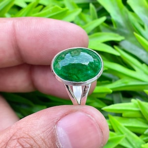 May include: A silver ring with a large, oval emerald-green gemstone. The gemstone is set in a polished silver band. The ring's design is simple and elegant. The ring is held against a backdrop of green foliage.