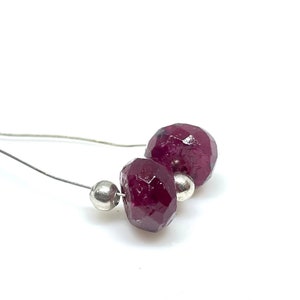 May include: Close-up of a pair of faceted, deep red gemstone beads strung on a thin, silver wire. Two small, silver beads are also strung on the wire. The gemstones have a geometric, multi-faceted cut. The background is white.