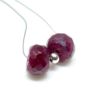 May include: Close-up of two faceted, deep red gemstone beads strung on a thin, light-colored wire. A small, silver-colored bead is between the two larger beads. The beads have a geometric, multi-faceted surface. The background is white.