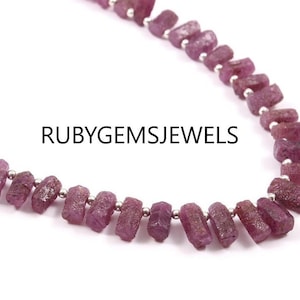 May include: A strand of raw ruby gemstone beads. The beads are a deep pinkish-red color and are strung on a silver-colored wire. The text "RUBYGEMSJEWELS" is displayed in black.