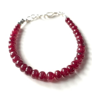 May include: A beaded bracelet featuring small, round, deep red beads. The beads are strung together with a silver-colored clasp. The bracelet is displayed against a white background, highlighting the color and texture of the beads.