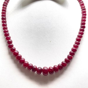 May include: A necklace featuring a strand of polished, deep red gemstones. The beads are evenly sized and strung together, creating a smooth, curved shape. The necklace is displayed against a white background, highlighting the color and shine of the gemstones.