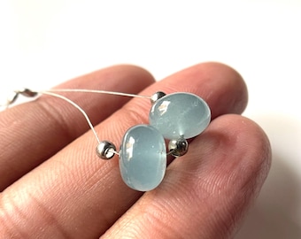 May include: Two light blue aquamarine gemstone beads with silver accents strung on a thin wire. The beads are oval-shaped and have a translucent appearance. The image is a close-up shot.