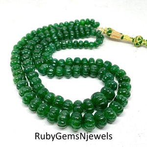 Op de afbeelding: Een ketting van gesneden, diepgroene kralen. De kralen hebben een rijke smaragdgroene kleur en hebben een geribbelde textuur. De ketting is geregen op een goudkleurig koord met groene accenten. De tekst "RubyGemsNjewels" is zichtbaar onderaan.