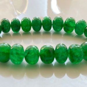 Könnte beinhalten: Ein Armband mit einer Kette aus leuchtend grünen, polierten Perlen. Die Perlen sind gleichmäßig groß und aufgereiht, wodurch eine kreisförmige Form entsteht. Das Armband wird auf einer reflektierenden Oberfläche präsentiert, die den Glanz der Perlen hervorhebt.