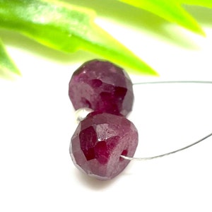 May include: Two faceted, deep red ruby beads strung on a thin, silver wire. The beads have a geometric cut, reflecting light. The background is white with a blurred green leaf.