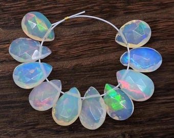 May include: Strung, faceted opal teardrop beads. The opals display iridescent flashes of color, including blue, green, and pink. The beads are strung on a white cord, and are displayed on a dark wood surface.