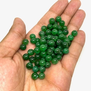 Puede incluir: Un puñado de pequeñas cuentas verdes redondas y pulidas. Las cuentas son de un color verde esmeralda vibrante y parecen estar hechas de un material liso y reflectante. Las cuentas están sueltas y dispersas en la palma de una mano.