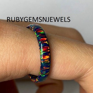 May include: A black opal ring with vibrant flashes of red, orange, and green. The ring is set against a light background. The text "RUBYGEMSNJEWELS" is visible in the image.