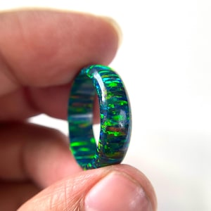 Puede incluir: Un anillo con una base oscura y motas iridiscentes vibrantes de color azul y verde por toda la banda. La superficie del anillo es lisa y pulida, mostrando un patrón multicolor y brillante que capta la luz a lo largo de su circunferencia.