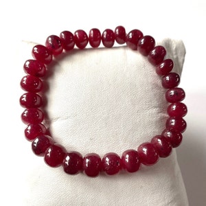 Pode incluir: Uma pulseira de contas com pedras preciosas polidas, de cor vermelha profunda. As contas redondas são enfiadas, criando um laço contínuo. A pulseira é exibida sobre um fundo branco, realçando a cor e a textura das pedras.