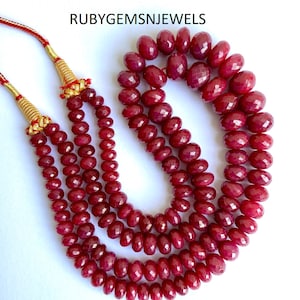 Puede incluir: Tres hilos de cuentas de piedras preciosas facetadas de color rojo intenso. Las cuentas están ensartadas con detalles dorados y un cordón rojo. El texto "RUBYGEMSNJEWELS" está impreso en negro.