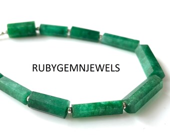 May include: A necklace featuring faceted, emerald green gemstone beads. The beads are rectangular with angled edges, strung together with small silver-colored connectors. The text "RUBYGEMNJEWELS" is printed in black.