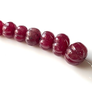 May include: A pair of earrings featuring a series of carved, deep red beads. The beads are spherical with a ribbed design, strung together on a silver post. The earrings are set against a white background.