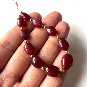 Peut inclure: Un fil de perles de rubis rouge foncé de forme ovale, avec des séparateurs dorés. Les perles sont enfilées sur un fil fin de couleur or, créant un composant de collier ou de bracelet. Les pierres précieuses sont polies et ont une finition brillante.