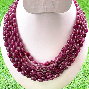 May include: A multi-strand necklace featuring oval-shaped, deep red gemstones. The necklace is displayed on a white mannequin bust against a backdrop of green grass. The gemstones have a polished finish and are strung together.
