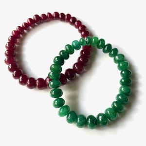 Può includere: Due braccialetti di perline. Uno è di colore rosso intenso, l'altro verde vibrante. Le perline sono rotonde e infilate insieme per formare una forma circolare. I braccialetti sono esposti su una superficie bianca.