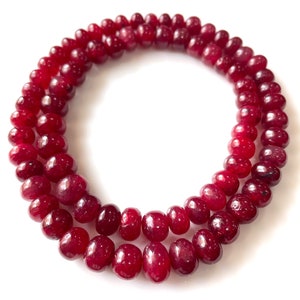 May include: Two strands of deep red gemstone beads, possibly ruby, are arranged in a circular pattern against a white background. The beads are smooth, rounded, and have a rich, saturated colour. Suitable for necklaces or bracelets, the beads are of high quality.