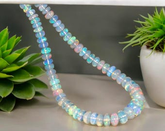 May include: A strand of iridescent opal beads. The beads are a mix of white, blue, pink, and green, creating a shimmering effect. The necklace is displayed on a white surface with green plants in the background. The beads are approximately 6mm in diameter.