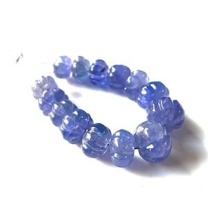 May include: A strand of carved, light blue gemstone beads. The beads have a floral design and are strung together. The beads are a light, translucent blue color and are on a white background.
