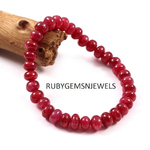 Könnte beinhalten: Ein rotes Edelsteinarmband mit abgerundeten Perlen. Das Armband besteht aus polierten, tiefroten Edelsteinen und ist aufgefädelt. Der Text "RUBYGEMSNJEWELS" ist auf dem Bild zu sehen. Das Armband wird auf einer weißen Oberfläche präsentiert.