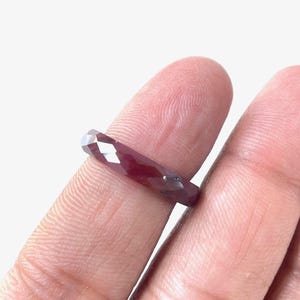 May include: A faceted, deep red gemstone ring. The ring is held between two fingers, showcasing its elongated, rectangular shape and the play of light across its cut surfaces. The gem's rich color is prominent.