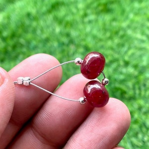 May include: A pair of earrings featuring two oval, deep red gemstones. The gemstones are connected by thin, silver-colored wires and small silver beads. The earrings are displayed against a blurred green background.