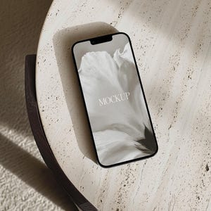 May include: A black smartphone with the word "MOCKUP" on the screen, resting on a light-colored, textured round table. The table has a dark brown curved edge. The background includes a soft, white rug and shadows.