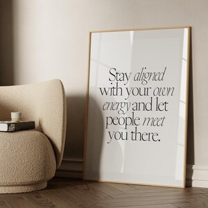 Peut inclure: Une affiche encadrée avec le texte "Stay aligned with your own energy and let people meet you there." est appuyée contre un mur. Un fauteuil beige avec un livre et une petite tasse se trouve à proximité. La pièce a une palette de couleurs neutres.