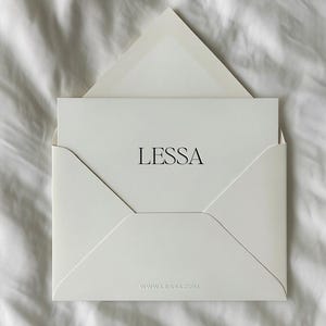 May include: A white envelope with a matching card inside, both bearing the name "LESSA" in a simple, elegant font. The website address "WWW.LESSA.COM" is printed at the bottom of the envelope. The items are displayed on a white, textured surface.