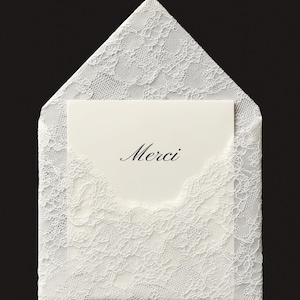 May include: A white lace envelope with a matching card inside. The card has the word "Merci" in elegant script. The envelope and card are set against a black background, highlighting the delicate lace pattern.