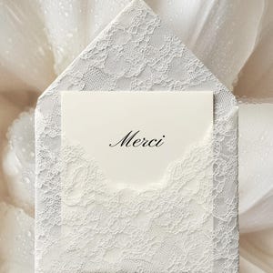 May include: A white lace-covered envelope with a matching card inside. The card has the word "Merci" printed on it in elegant script. The envelope and card are set against a soft, white background.