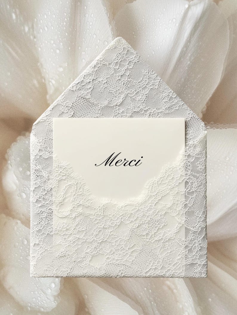 May include: A white lace-covered envelope with a matching card inside. The card has the word "Merci" printed on it in elegant script. The envelope and card are set against a soft, white background.