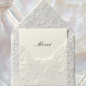May include: A white lace envelope with a matching card inside. The card has the word "Merci" in elegant script. The envelope and card are set against a pearlescent background.