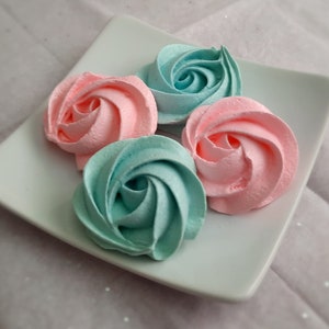 May include: Four pastel-colored meringue cookies shaped like roses on a square white plate. Two are pink, and two are teal. The plate sits on a light pink, textured surface.
