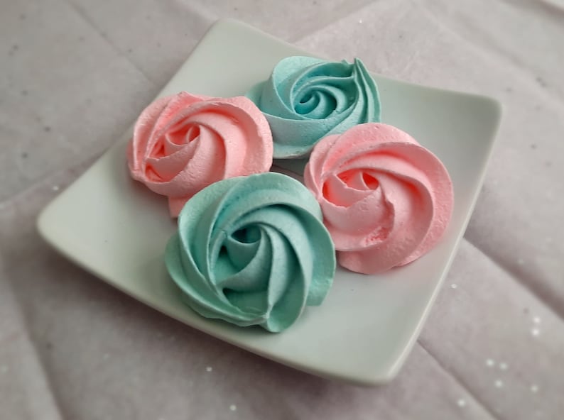 May include: Four pastel-colored meringue cookies shaped like roses on a square white plate. Two are pink, and two are teal. The plate sits on a light pink, textured surface.
