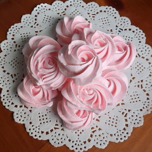 May include: A cluster of pink, rose-shaped meringues arranged on a white, lacy paper doily. The meringues have a light pink hue and are swirled to resemble rose petals. The doily sits on a wooden surface.