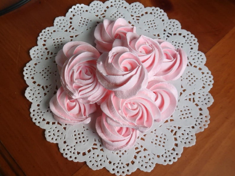 May include: A cluster of pink, rose-shaped meringues arranged on a white, lacy paper doily. The meringues have a light pink hue and are swirled to resemble rose petals. The doily sits on a wooden surface.