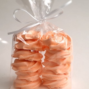 May include: A clear cellophane bag tied with a white ribbon, filled with peach-colored meringue cookies. The cookies are stacked in two columns, showcasing their swirled design. The background is a plain white surface.
