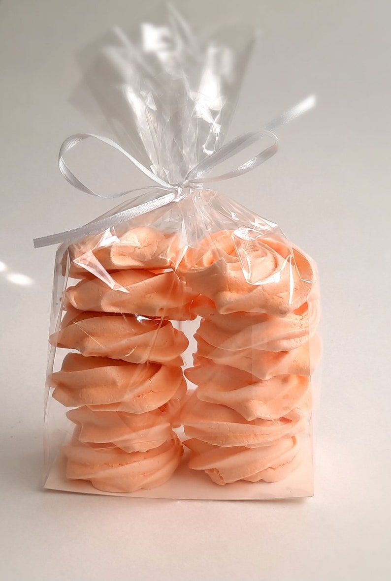 May include: A clear cellophane bag tied with a white ribbon, filled with peach-colored meringue cookies. The cookies are stacked in two columns, showcasing their swirled design. The background is a plain white surface.