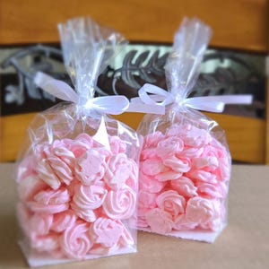 May include: Two clear cellophane bags filled with pink meringue cookies tied with white ribbon. The cookies are in various shapes, including hearts and swirls. The background is a blurred wooden surface.