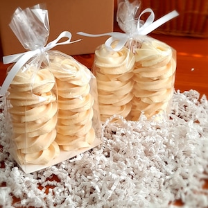 May include: Two clear cellophane bags tied with white ribbon, each filled with stacks of pale yellow, swirled cookies. The cookies sit on a bed of white shredded paper. A brown cardboard box and a wicker basket are in the background.
