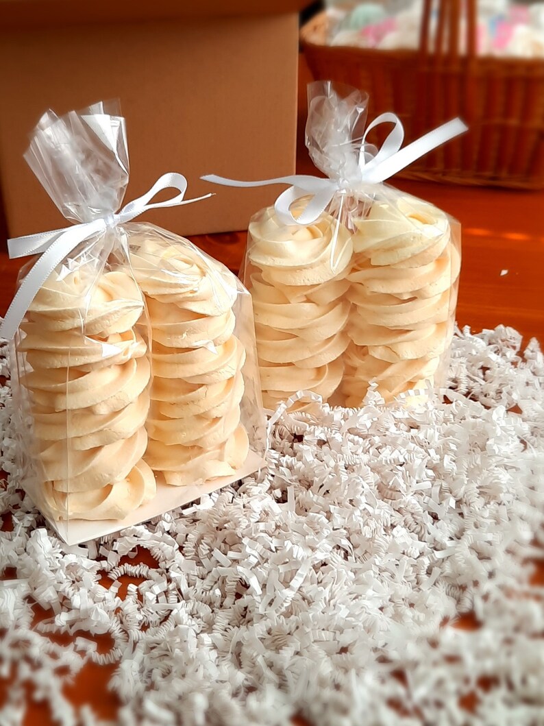 May include: Two clear cellophane bags tied with white ribbon, each filled with stacks of pale yellow, swirled cookies. The cookies sit on a bed of white shredded paper. A brown cardboard box and a wicker basket are in the background.