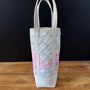 May include: A quilted wine tote bag with a cream-colored base and handles. The body of the bag features a patchwork design in pink, blue, and cream. The bag has a rectangular shape and is designed to hold a single bottle.