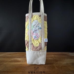May include: A quilted wine bottle tote with a patchwork design. The tote features a cream-colored base and handles, with a central panel of yellow, brown, and pastel-colored fabric. A small white tag is attached to the side.